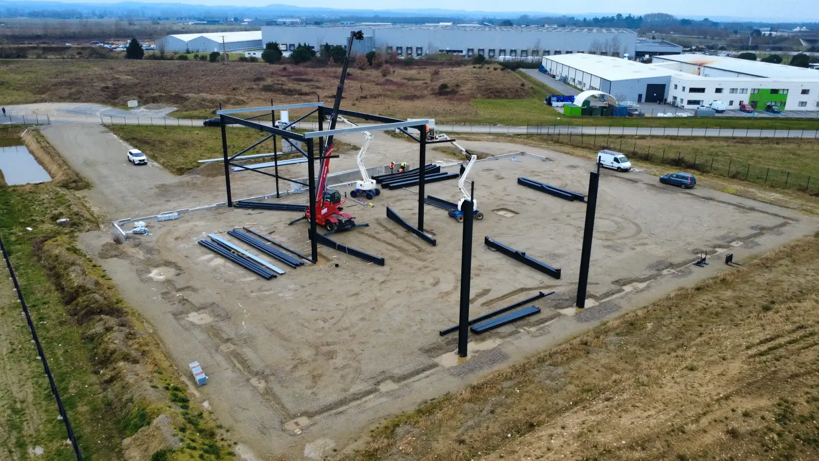 Vue aérienne d’un chantier de bâtiment d’activité : poteaux et poutres métalliques en cours de montage, grues et nacelles sur un terrain nu