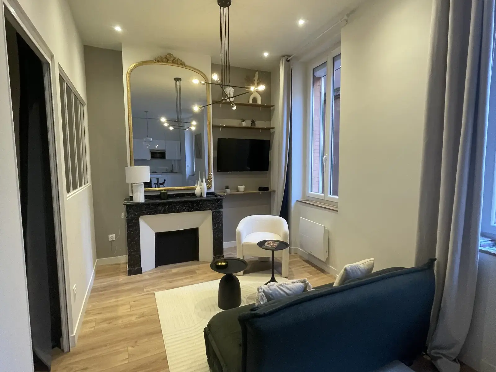 Même pièce rénovée : cheminée en marbre noir, grand miroir doré, spots au plafond, parquet clair et canapé vert