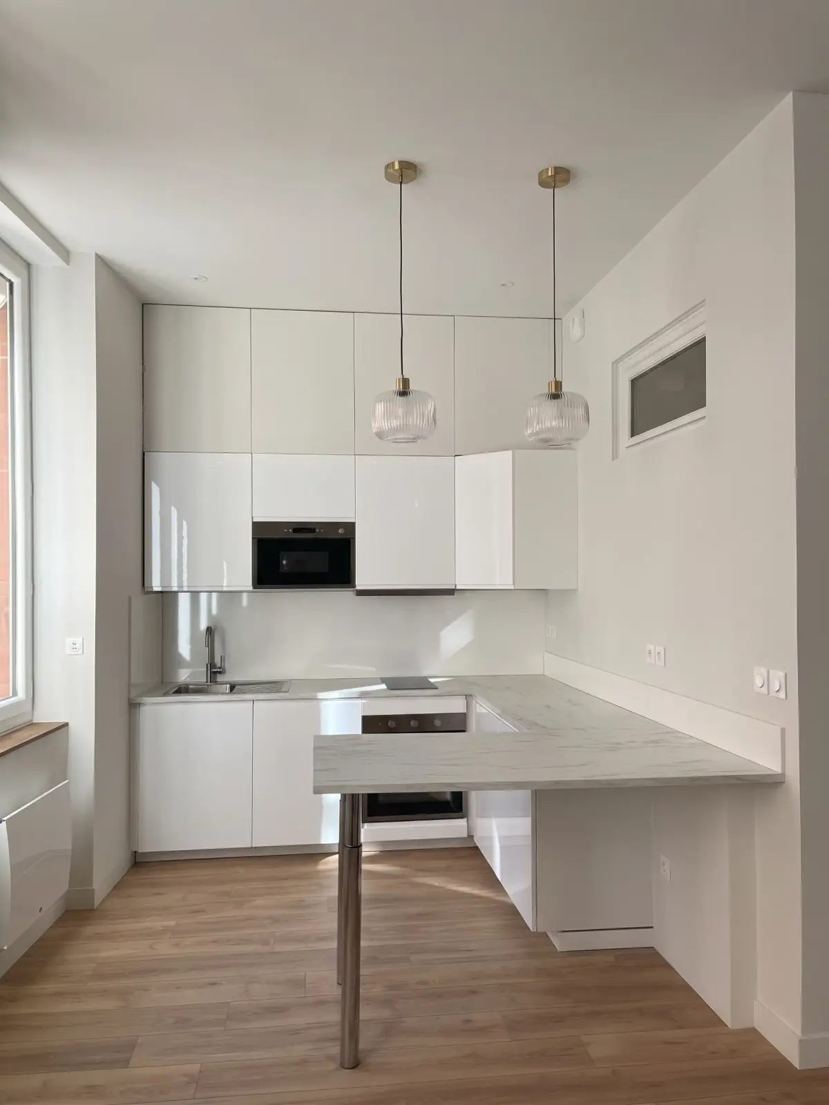 Cuisine blanche sous deux suspensions, avec péninsule à plan de travail en marbre et parquet clair