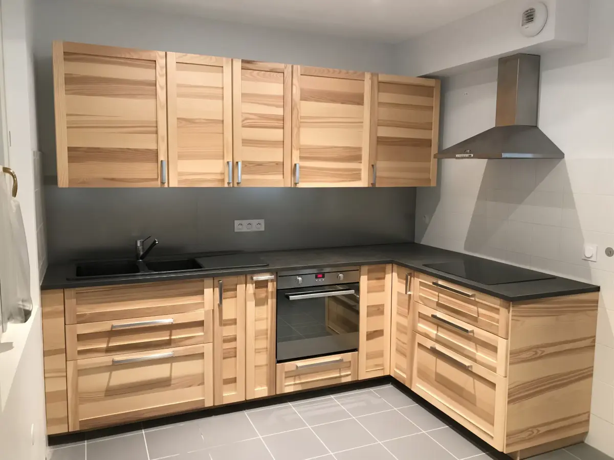 Cuisine en L aux façades en bois clair, plan de travail noir et hotte inox
