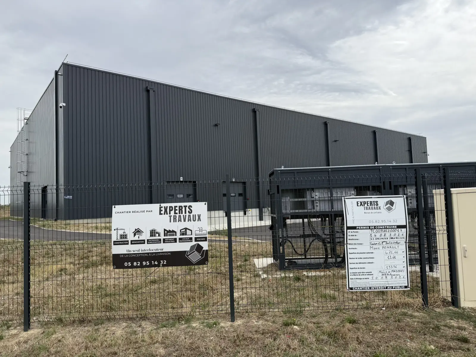 Bâtiment industriel à bardage noir derrière une clôture, avec le panneau de chantier Experts Travaux et le panneau du permis de construire
